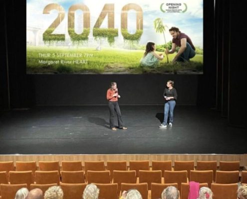 Startbeeld documentaire 2040 op groot filmscherm in een verlichte filmzaal met mensen op stoelen kijkend naar het podium en het filmscherm. Twee mensen op het podium voor het filmscherm die in gesprek zijn over de film die vertoond gaat worden.